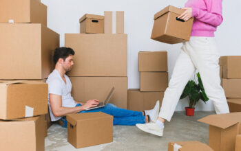 Crop woman arranging carton boxes during relocation with boyfriend using laptop on floor